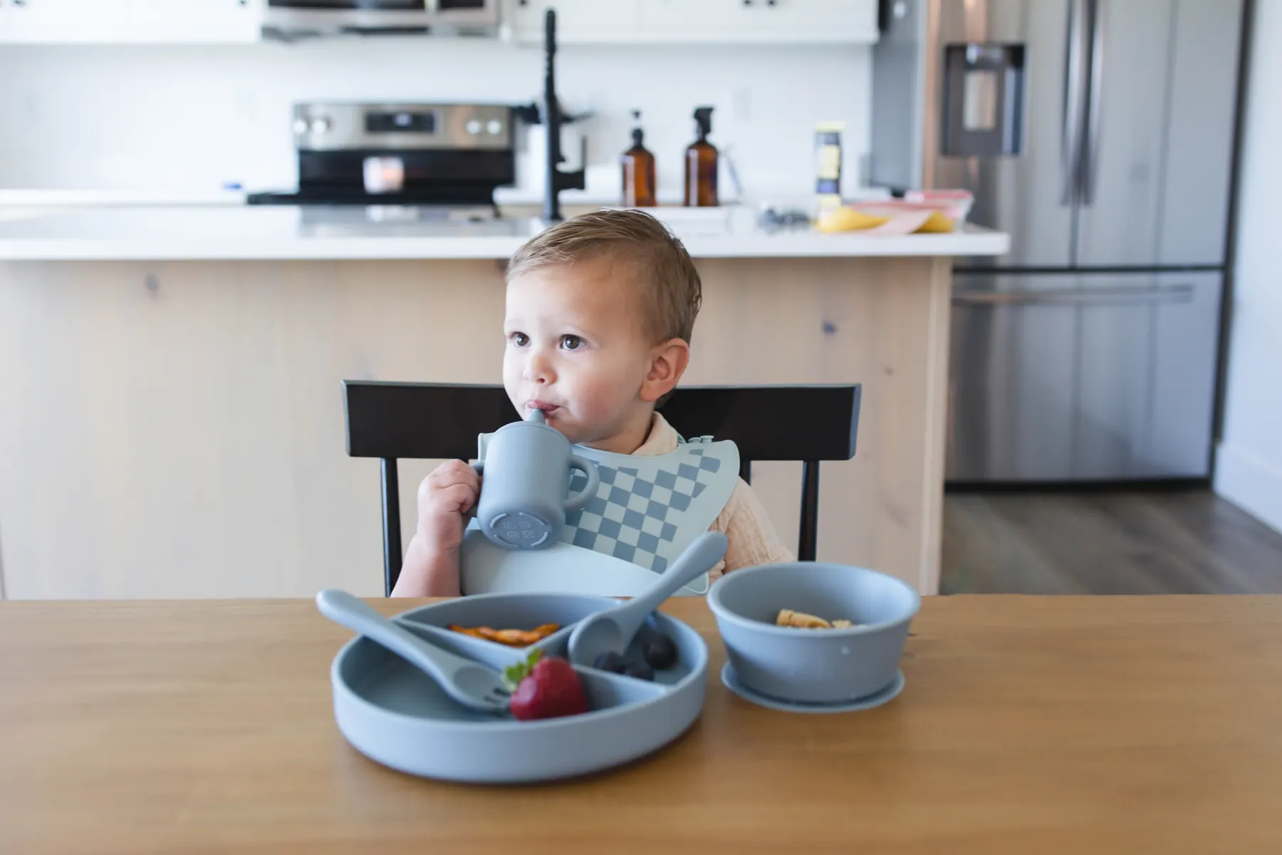 A toddler eating at a kitchen table wearing a light blue checkered KAYA silicone bib