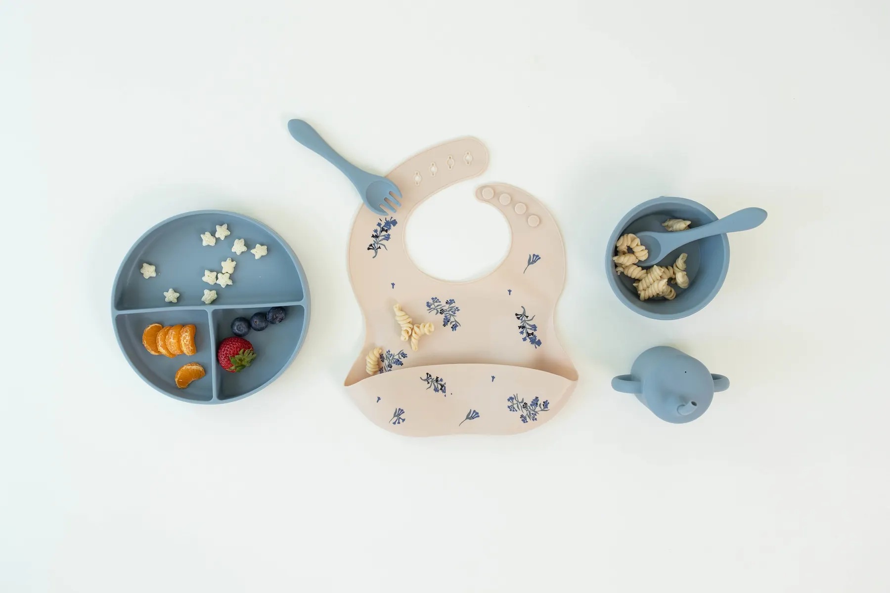 A cream KAYA silicone bib with a floral print laid flat beside blue plates and bowls of food