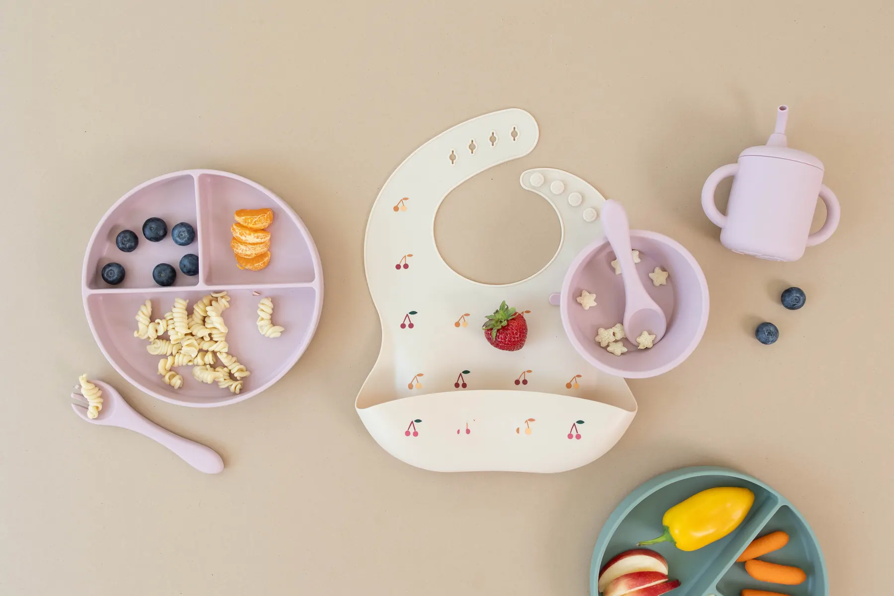 A cream KAYA silicone bib with a cherry print laid out with a divided plate of fruit and a small cup