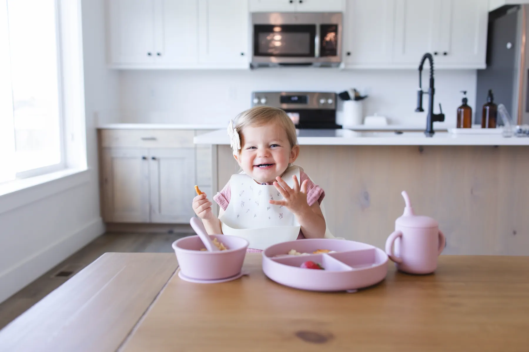 A toddler smiling at a kitchen table wearing an ivory KAYA silicone bib with a wildflower print