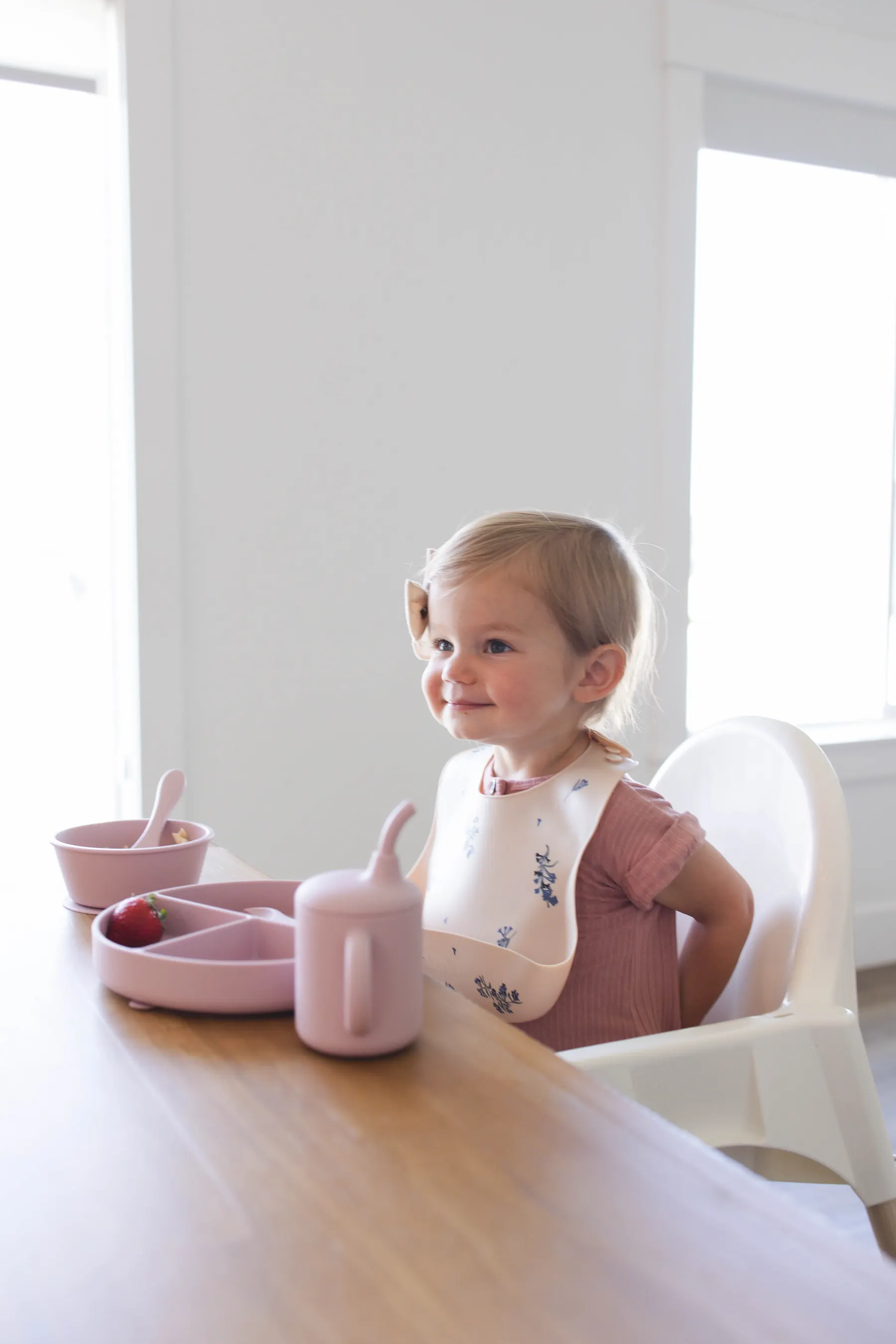 A toddler sitting at a table wearing a blush KAYA silicone bib with a lilac flower print