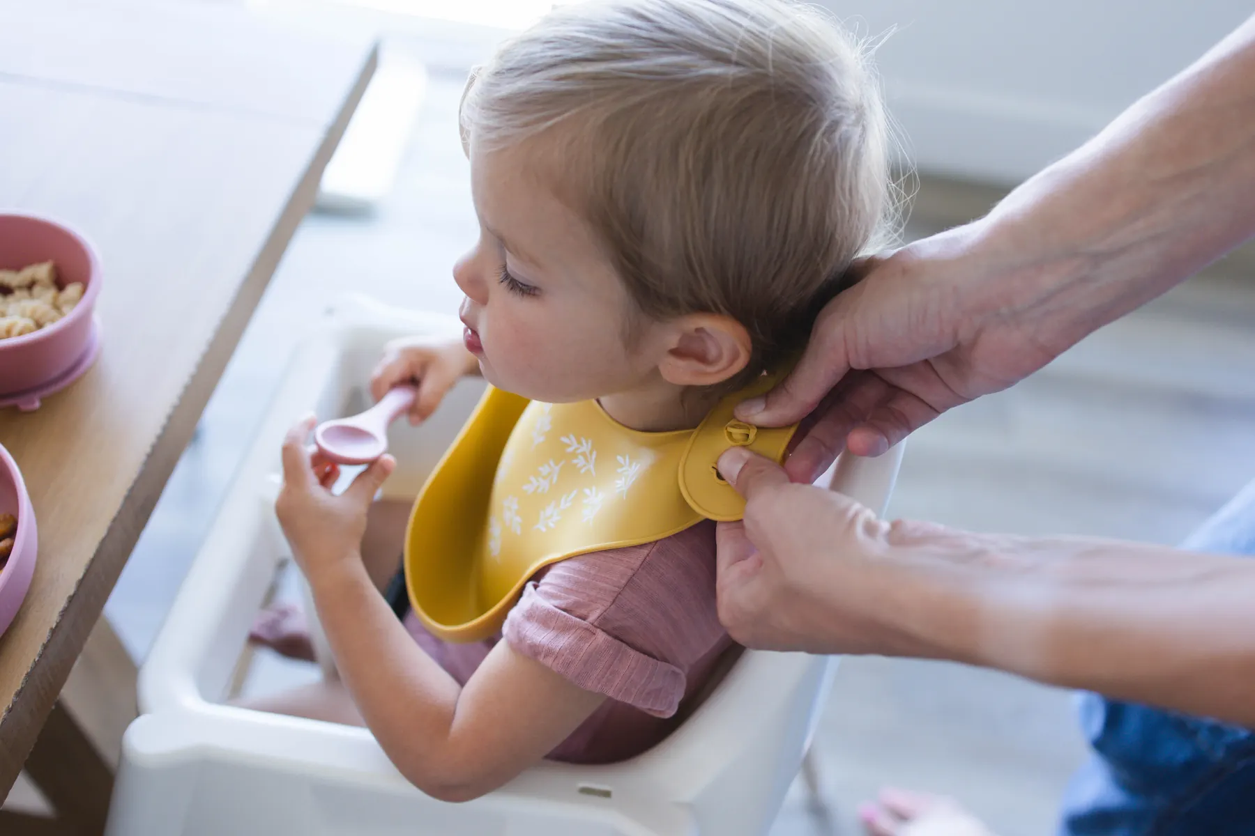 A parent fastening the snap neckband of a yellow KAYA silicone bib on a baby in a high chair