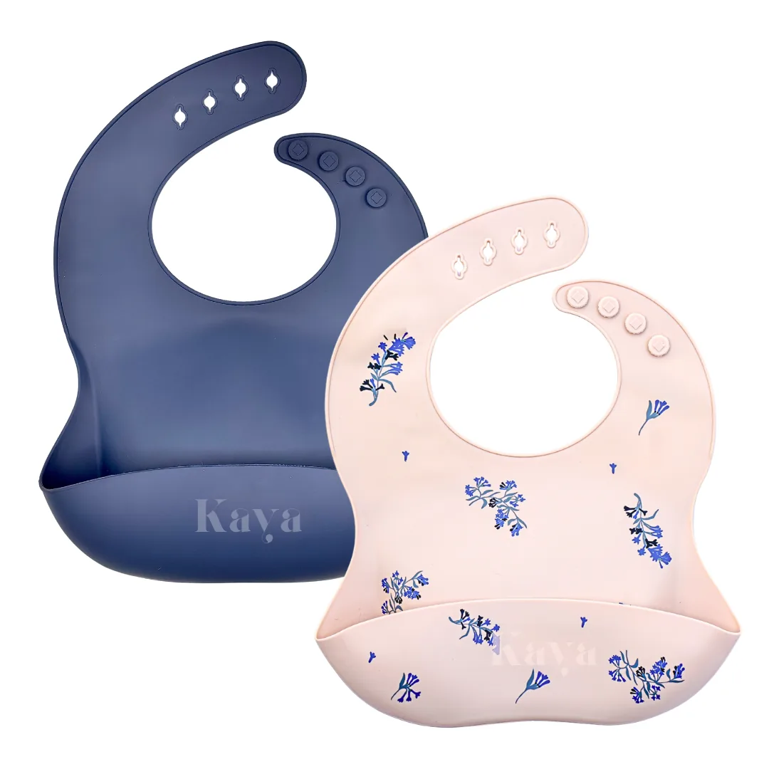 KAYA 2-pack of silicone baby bibs: a deep navy blue bib and a blush pink bib with a blue and lilac flower print, each with a food-catcher pocket and snap neck