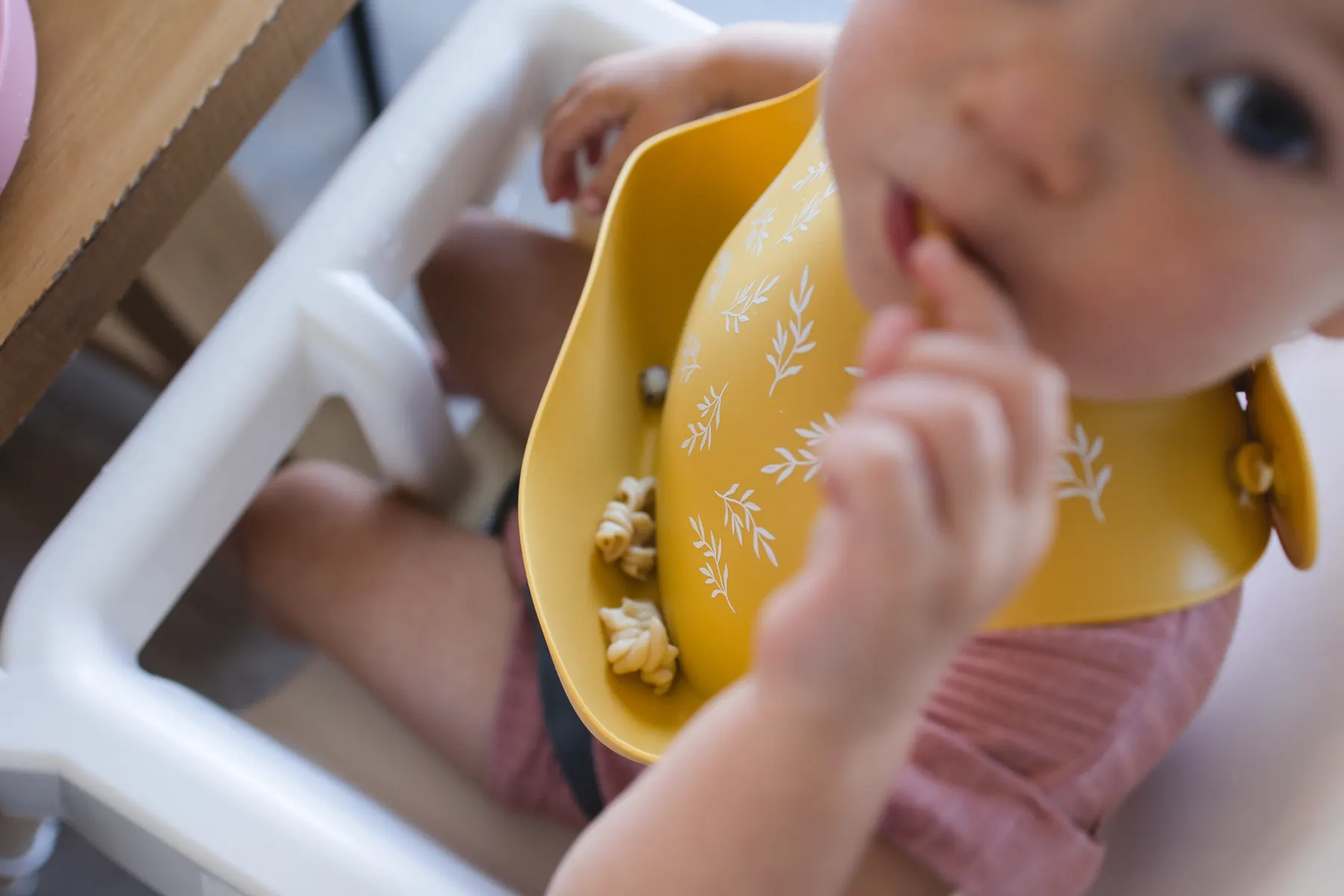 A baby in a high chair wearing a yellow KAYA silicone bib, eating finger food with their hands
