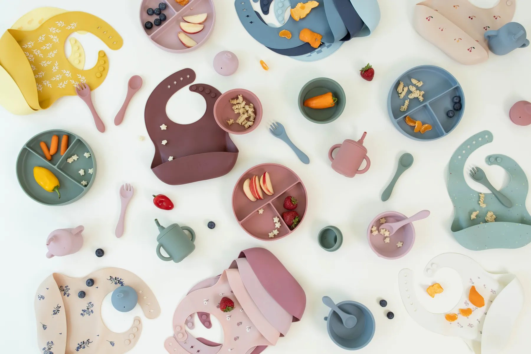 KAYA silicone bibs in several colors laid flat among plates of fruit and snacks