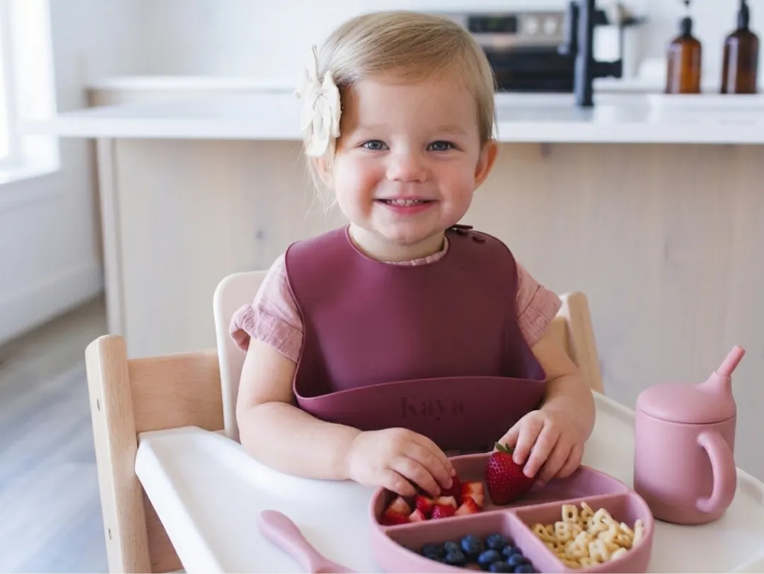 A smiling toddler in a high chair wearing the deep mauve KAYA silicone bib, with berries and crackers on the tray