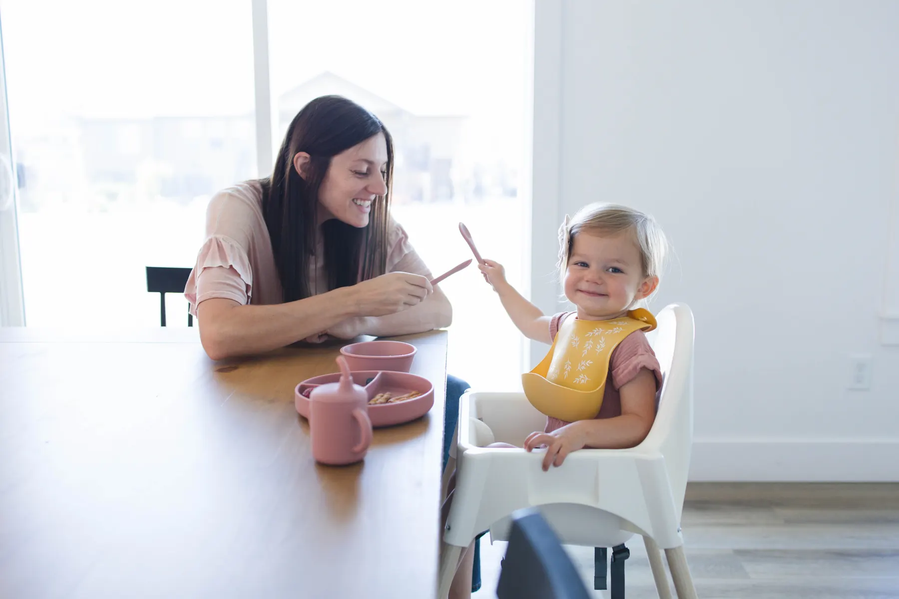 A parent and baby sharing a meal at the kitchen table, the baby wearing a yellow KAYA silicone bib
