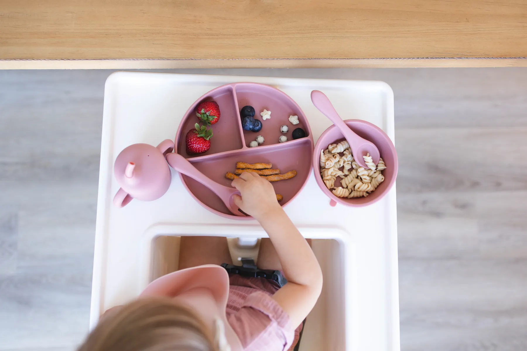 Overhead view of a high chair tray with a divided plate of berries and crackers and a bowl of cereal